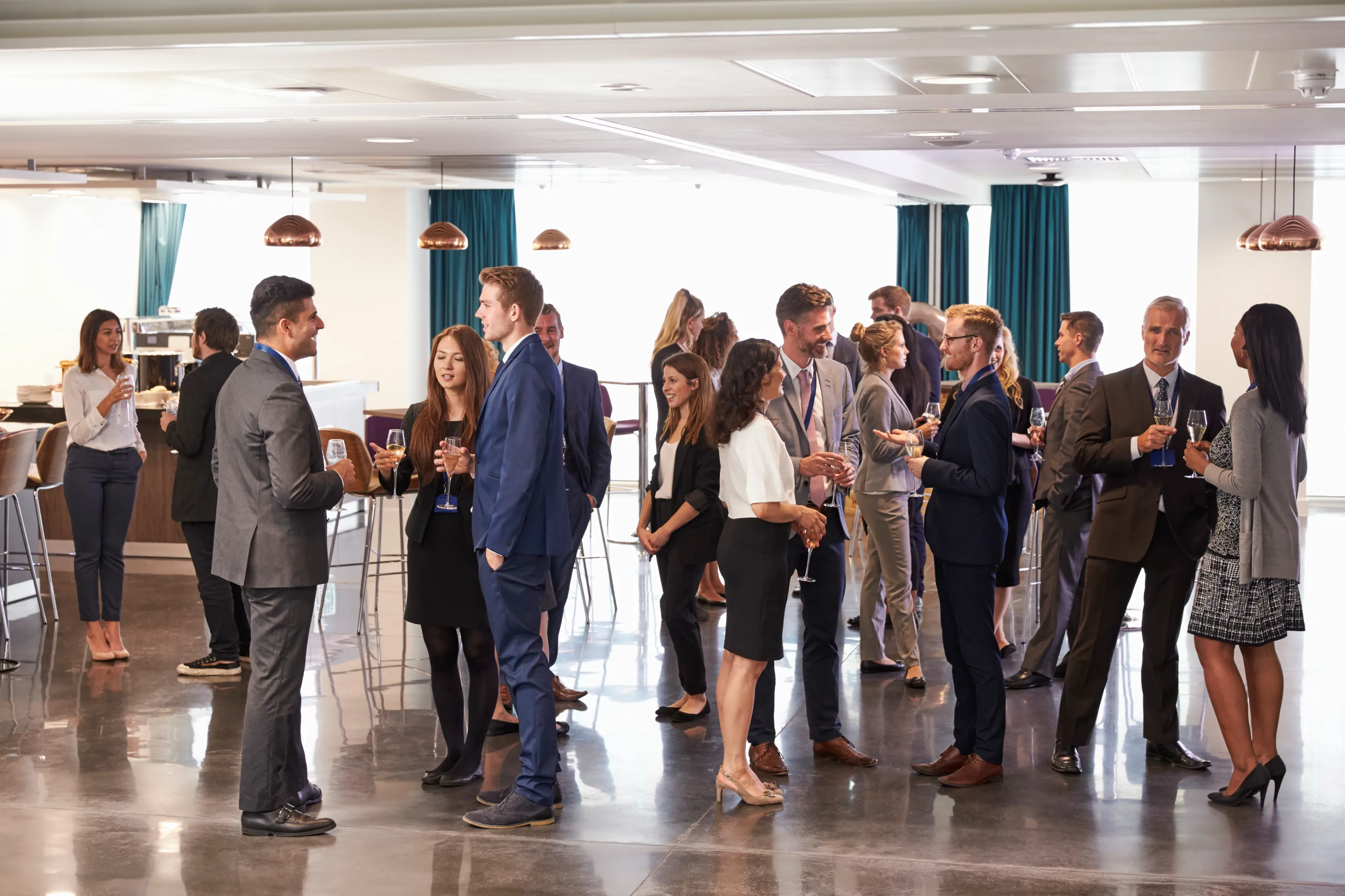People in business attire socializing and holding drinks at a networking event in a modern indoor setting.