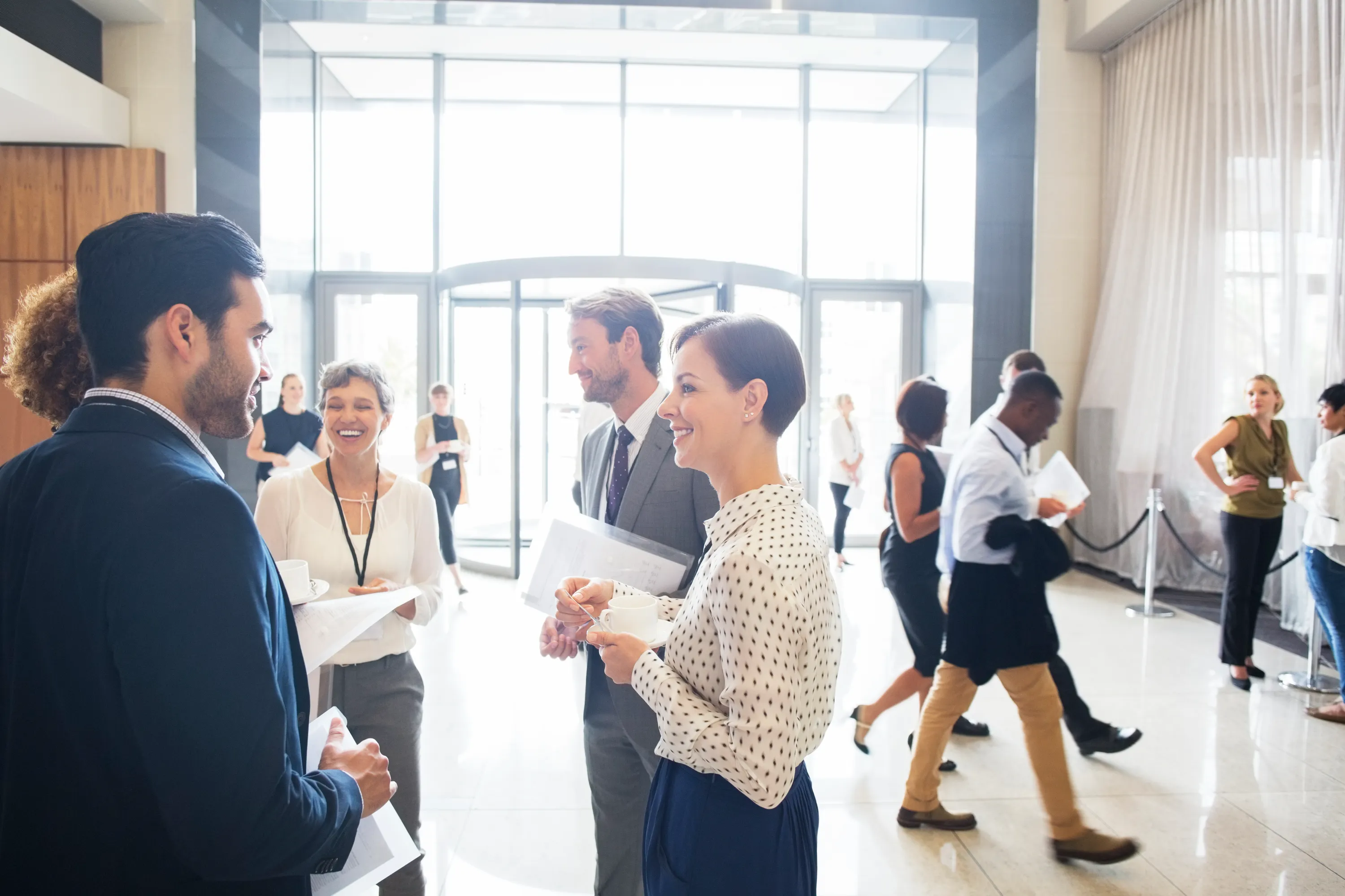 A group of professionals in business attire conversing and holding documents in a bright, open office lobby. Others are walking in the background.