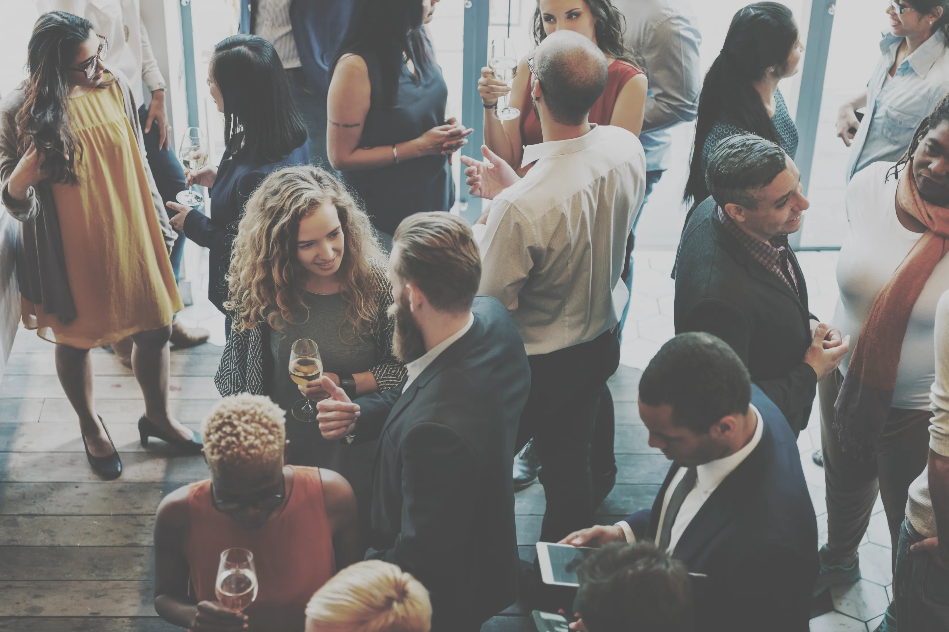 People socialize at a crowded indoor gathering. They are dressed in business or semi-formal attire, holding drinks, and engaging in conversation.