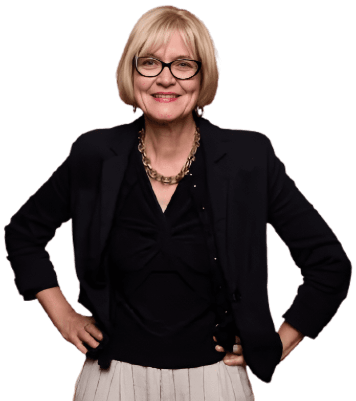 A woman with short blonde hair and glasses, wearing a black jacket and top with a pleated skirt. She is smiling with hands on hips.
