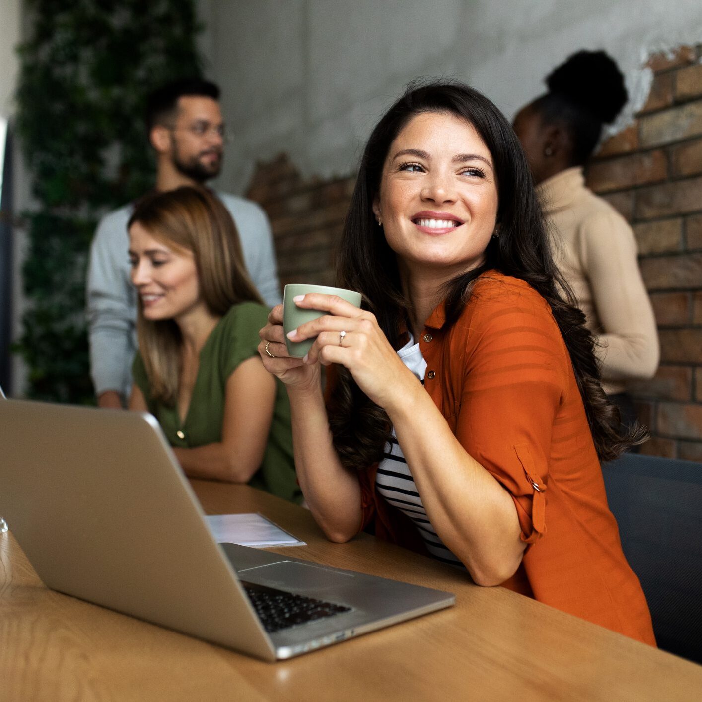 A smiling woman with a cup, sitting at a table with a laptop and people in the background.
