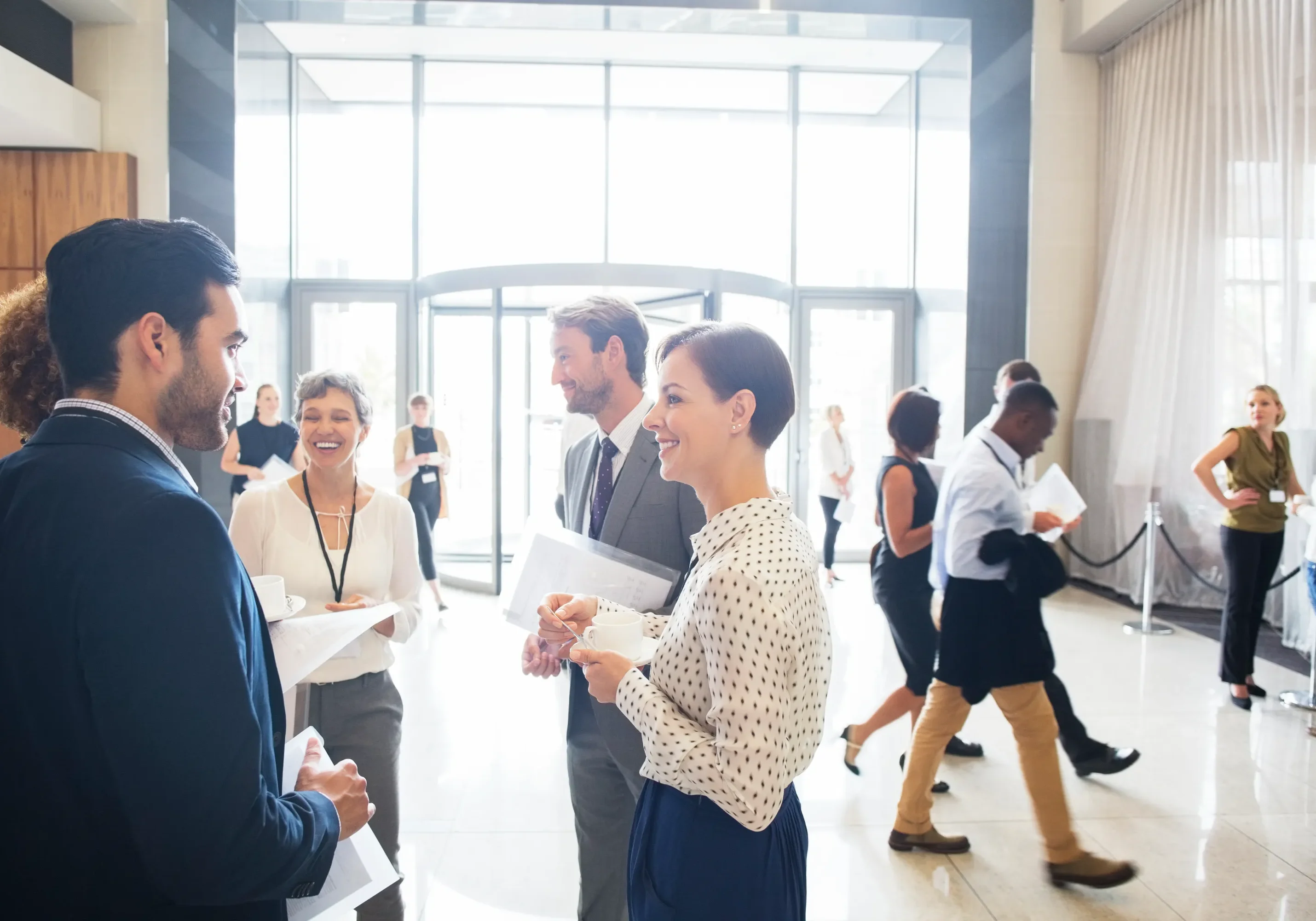A group of professionals in business attire conversing and holding documents in a bright, open office lobby. Others are walking in the background.