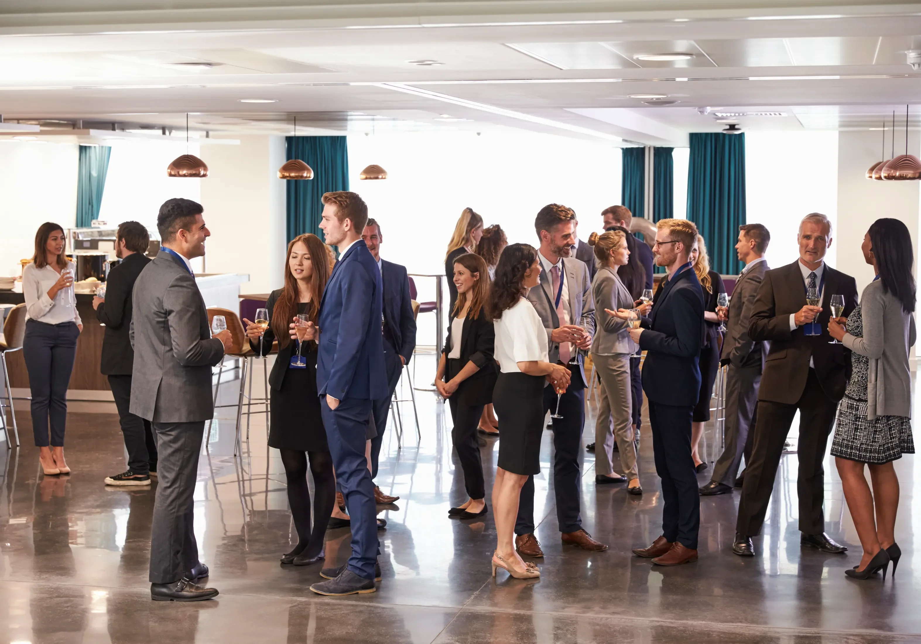 People in business attire socializing and holding drinks at a networking event in a modern indoor setting.