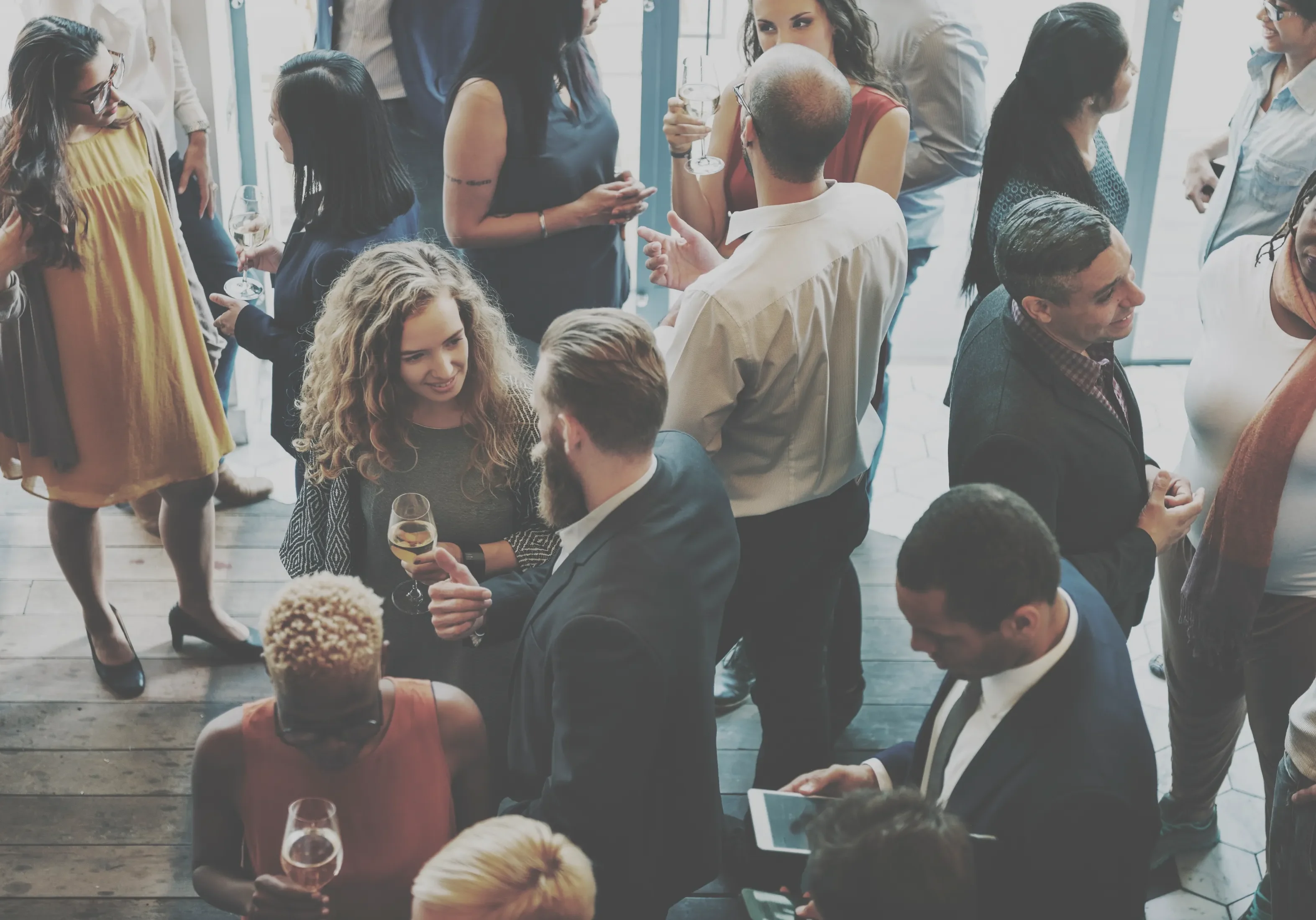 People socialize at a crowded indoor gathering. They are dressed in business or semi-formal attire, holding drinks, and engaging in conversation.